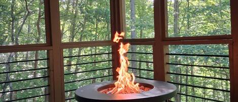 Un pozo de fuego circular al aire libre con llamas, rodeado por una terraza de madera y amplias ventanas con vistas a un área boscosa.