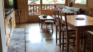 A cozy cabin interior with a wooden dining table, chairs, a small kitchen area, and a large window offering a view of the outdoors.