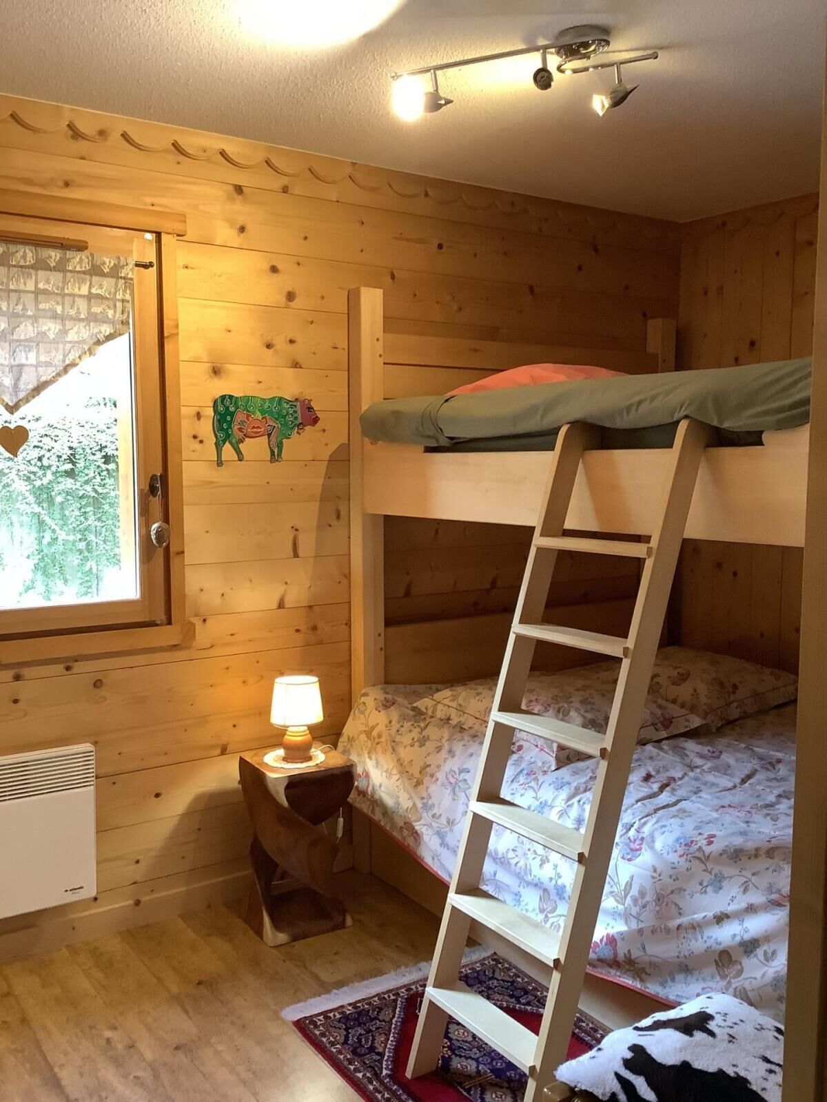 A cozy cabin room with a bunk bed, a small table with a lamp, a heater, and a window with a view of trees.