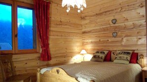 A wooden cabin bedroom with a bed, a chair, a window with curtains, and a lamp. - Ground floor, terrace, parking, tv, ski locker, 39m², Le Grand Bornand (Le Grand-Bornand)