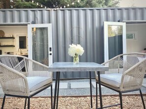 Un espace extérieur aménagé avec deux fauteuils en osier et une table, un vase rempli de fleurs blanches, et un mur métallique à rayures en arrière-plan.