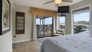 A bedroom with a large bed, a ceiling fan, a TV, and a balcony with outdoor furniture. - Waterfront 2BR/2BA Condo, Walk-Out Deck to Lake, Pools & Hot Tub (Village of Four Seasons)
