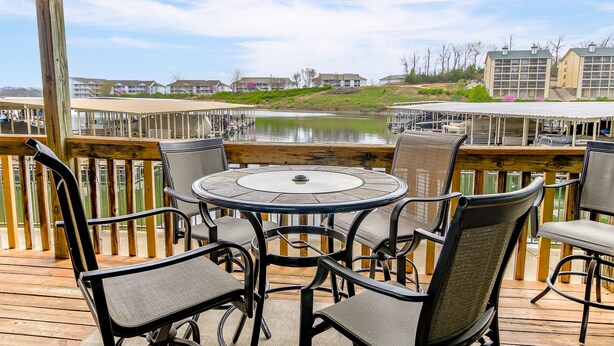 A covered outdoor area with a round table and chairs, overlooking a body of water and buildings in the background. - Waterfront 2BR/2BA Condo, Walk-Out Deck to Lake, Pools & Hot Tub (Village of Four Seasons)
