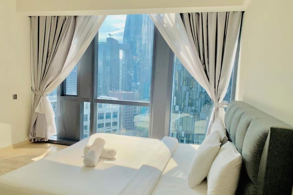 A modern hotel room with a large bed, white linens, and a view of a cityscape through floor-to-ceiling windows.