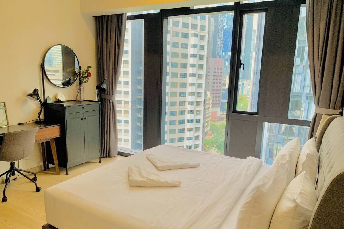 A hotel room with a bed, a desk, a chair, a mirror, and a view of tall buildings.