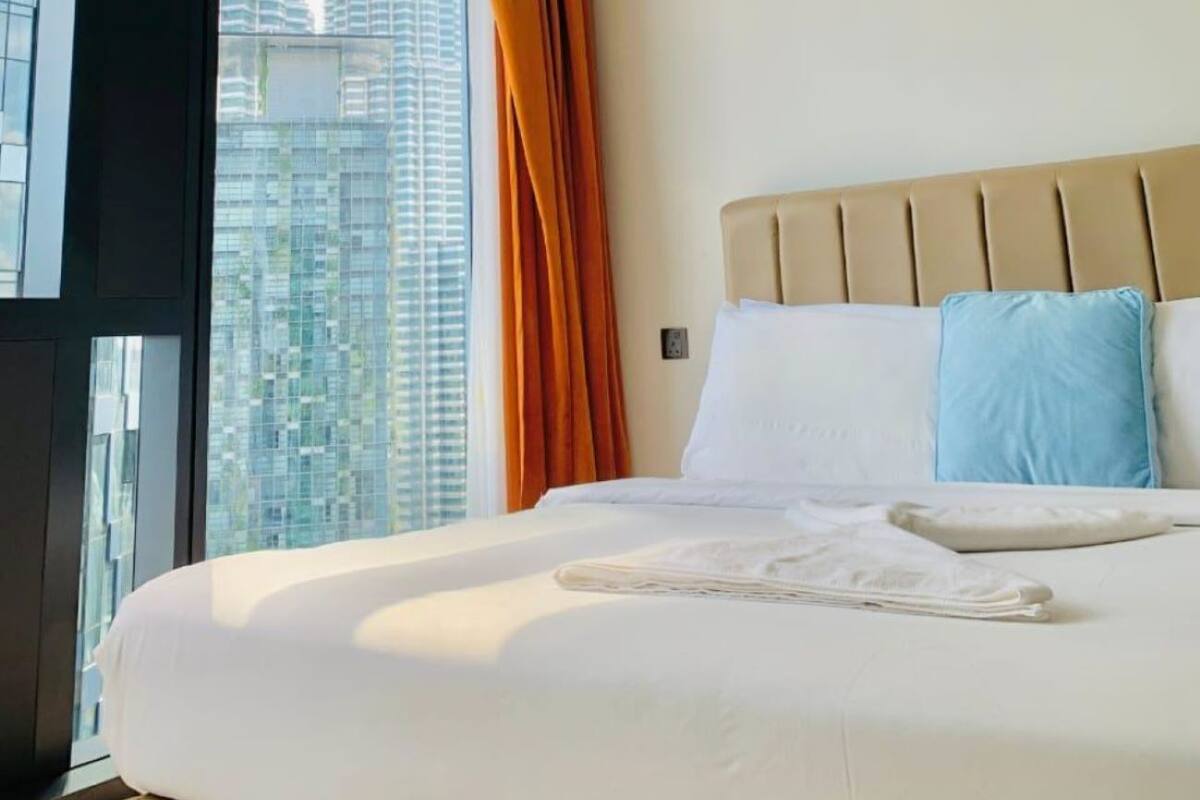 A hotel room with a large bed, a view of tall buildings, and a window with orange curtains.
