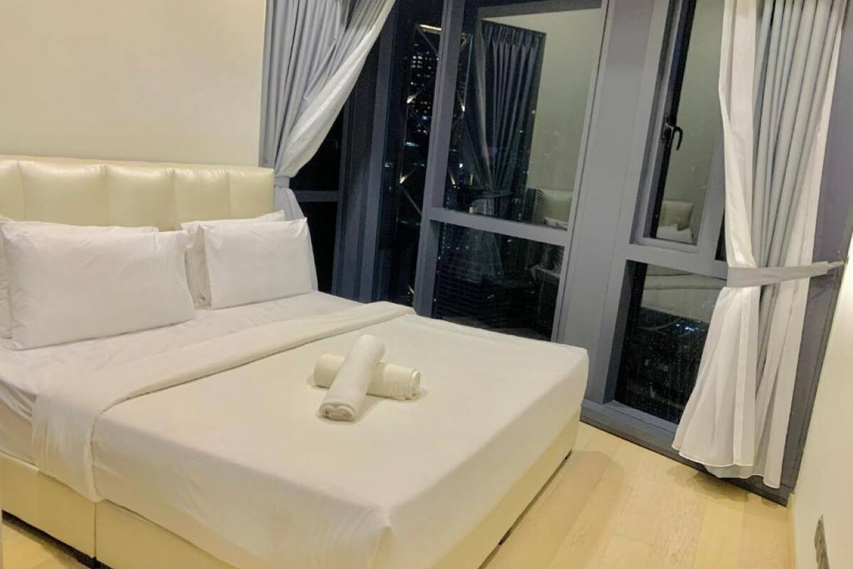 A neatly made bed with white linens and two rolled towels, a window with curtains, and a cityscape view at night.