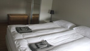 Una cama bien hecha con sábanas blancas y dos toallas dobladas, una lámpara de noche y una cesta de mimbre en una mesita de noche.