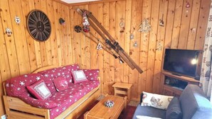 Un salon chaleureux avec un mur en bois, un canapé rouge à motifs blancs, un fauteuil bleu, une table basse en bois et un téléviseur.