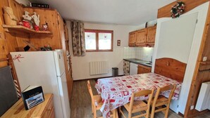 Une cuisine de taille réduite, dotée de meubles de rangement en bois, d’une table à manger avec des chaises, d’un réfrigérateur et d’une fenêtre avec des rideaux.