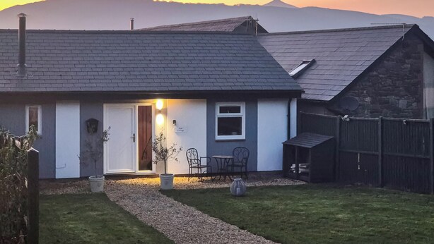 Una casa con fachada blanca y techo oscuro, un pequeño jardín delantero con un sendero, una silla y una mesa afuera.