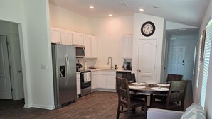 A modern kitchen with white cabinets, a stainless steel refrigerator, and a microwave. There is a dining area with a wooden table and chairs, and a gray sofa with a patterned throw pillow. - Charming 3-bedroom house with WiFi, AC in terrific Palm Coast (Palm Coast)