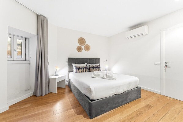 A modern bedroom with a grey upholstered bed, a bedside table with a lamp, a wall-mounted air conditioner, and a door with a handle.