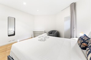 A hotel room with a large bed, a bedside table, a chair, a desk, a mirror, and a window with curtains.