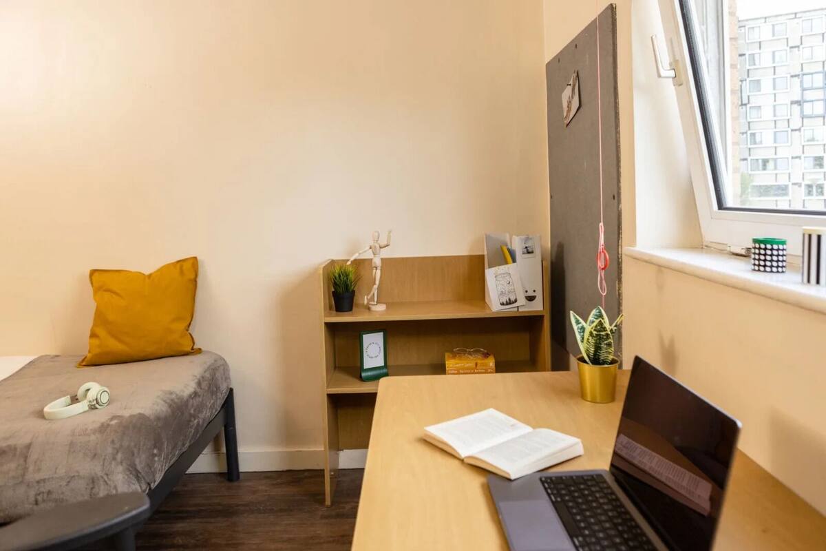 Una stanza piccola e ordinata con un letto, una scrivania con un portatile, una libreria e una finestra vista edifici.