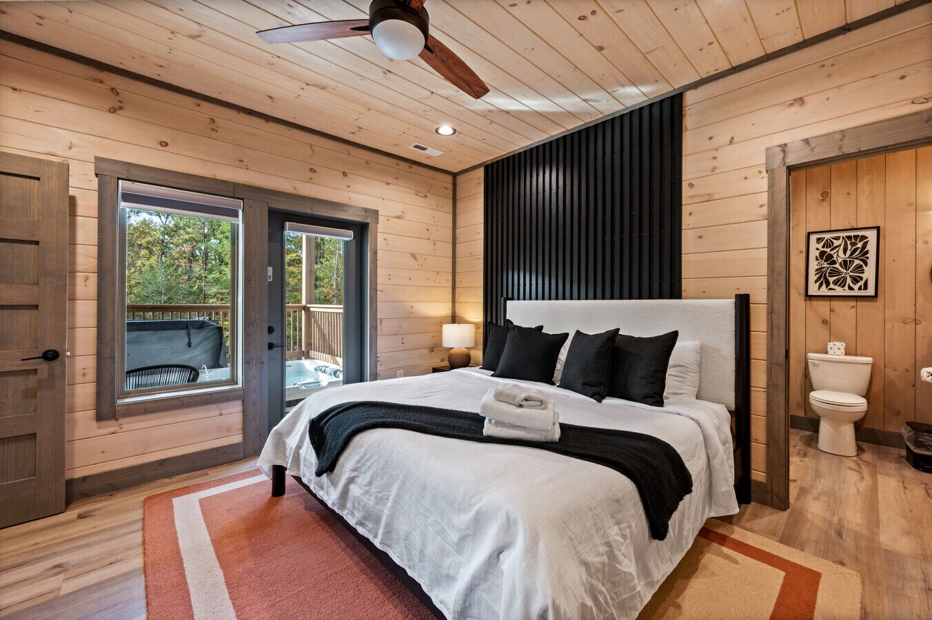Un dormitorio con una cama grande, un ventilador de techo, una ventana con vistas a árboles y un suelo de madera.