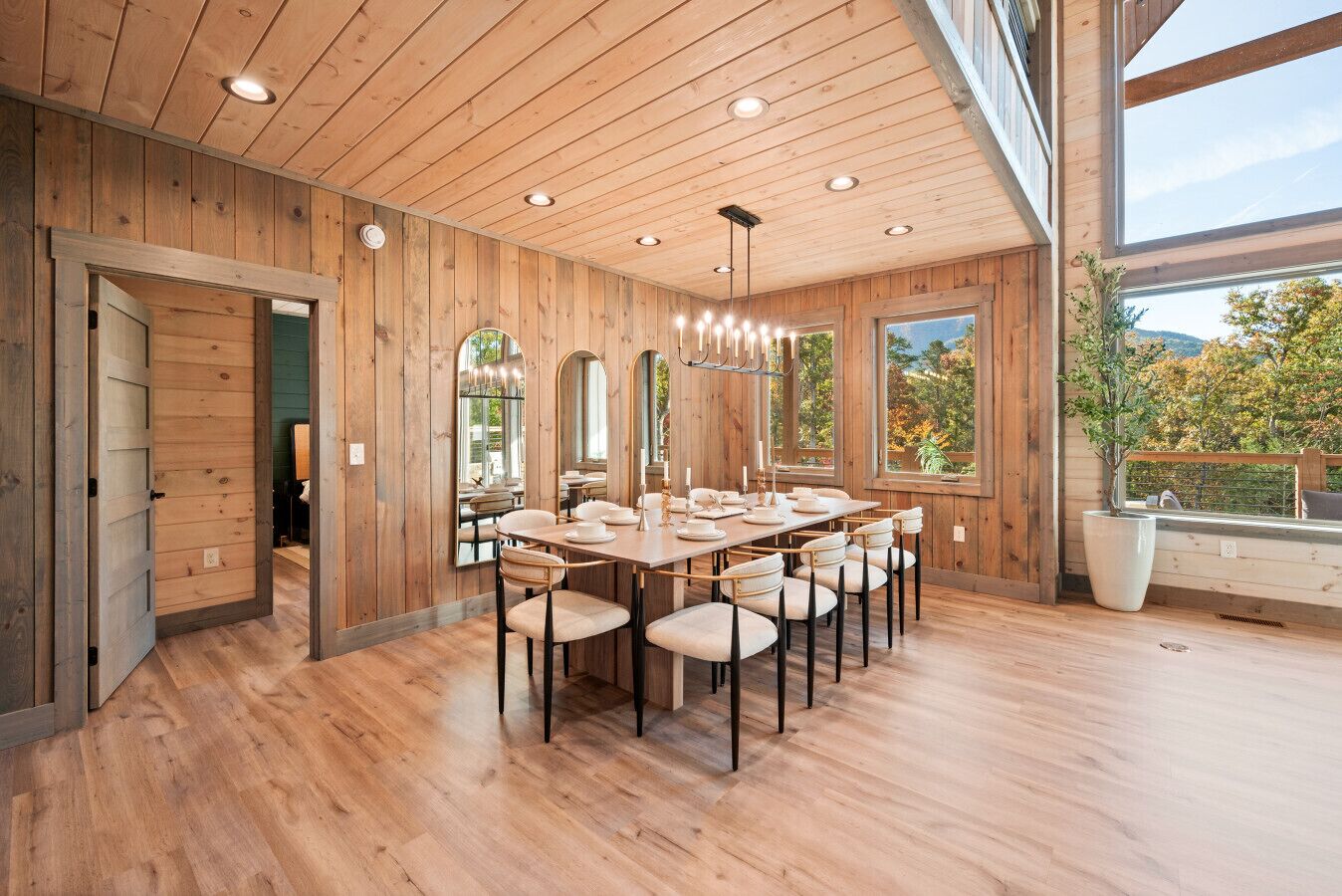 A spacious dining area with a long wooden table and chairs, large windows offering a view of the outdoors, and a wooden ceiling with recessed lighting.