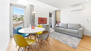 A modern living room with a grey sofa, a round table with chairs, a TV, and a view of a cityscape through a large window. - GuestReady - Riverside Retreat with Terrace Views (Vila Nova de Gaia)