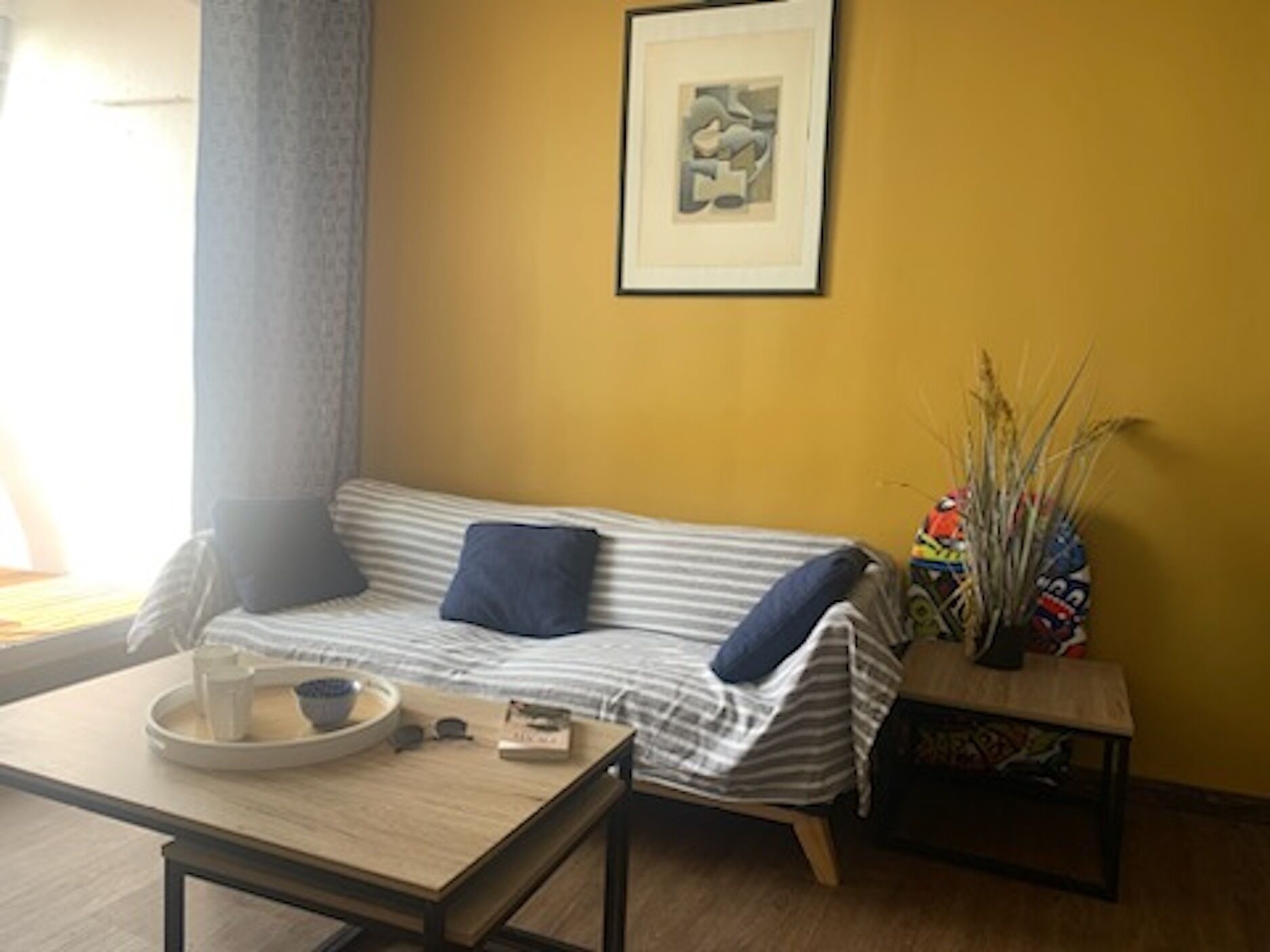Ett mysigt vardagsrum med en randig soffa, ett litet bord med en skål och böcker, ett inramat abstrakt konstverk på väggen och en prydnadsväxt på ett sidobord.