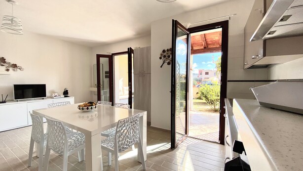 A modern kitchen with a large island, white cabinetry, and a dining area with a white table and chairs. - Villasimius, Vacation House, with private garden, Casagora (Sardegna)