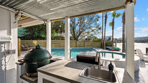 An outdoor kitchen with a grill, a sink, and a view of a pool and palm trees. - Captain's Cove 4BR 3Bath  Water View, Pickleball, Private Beach, Heated Pool! (Pensacola)