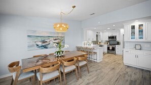A modern kitchen with white cabinets, a central island, and a dining area with a wooden table and chairs. - Captain's Cove 4BR 3Bath  Water View, Pickleball, Private Beach, Heated Pool! (Pensacola)