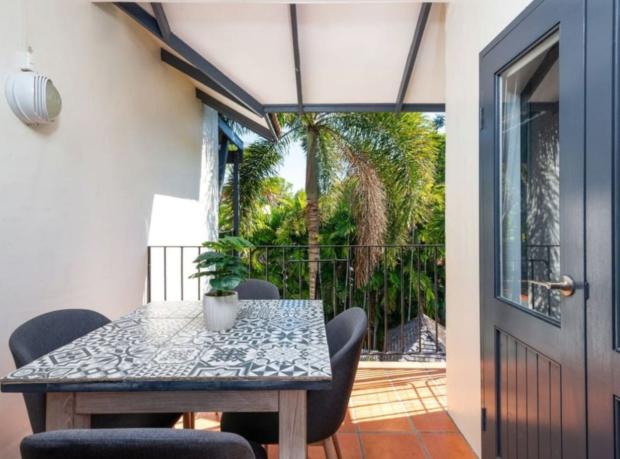 A patio with a table and chairs, a potted plant, and a view of a garden with palm trees.