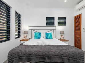 A modern bedroom with a large bed, two bedside tables with lamps, a headboard, and a window with blinds. - Holiday-Apartment Ocean Sound (Port Douglas)