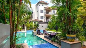 Un área de piscina con sillones reclinables, un edificio blanco con balcones y exuberante vegetación tropical.