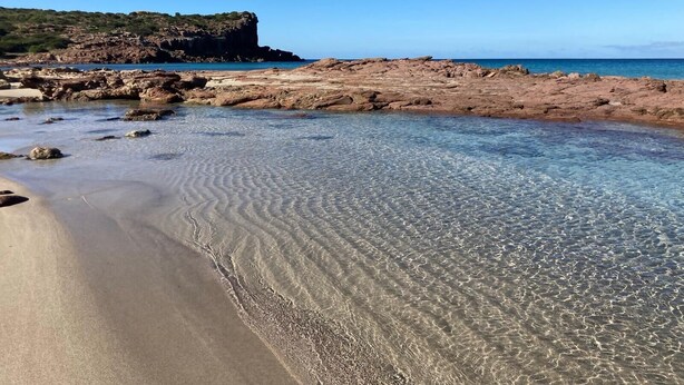 Une plage avec une eau claire et peu profonde, un terrain rocheux et une falaise qui se dresse au loin.