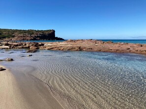 Una playa con aguas cristalinas y poco profundas, terreno rocoso y un acantilado distante.