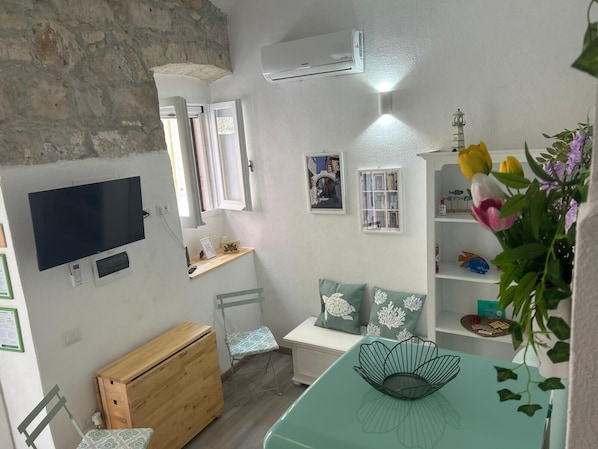 Una habitación con televisión, un escritorio pequeño, una silla, un armario de madera, una mesa verde y una estantería con flores.