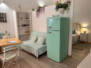 Una habitación compacta con un refrigerador verde azulado, una mesa de comedor de madera con sillas, un sofá, una estantería y una mesita de noche con lámpara.