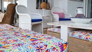 A room with a bed, two white wicker chairs with blue cushions, a small table with a white pitcher, and a colorful abstract painting on the wall.