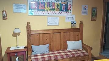Un sofà de fusta amb coixinets a viqueta, una petita taula de nit amb una làmpada, i una paret decorada amb obres d'art i diplomes.