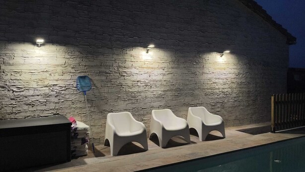 Un área junto a la piscina con un muro de piedra, tres sillas reclinables blancas y una pequeña mesa con un objeto azul.