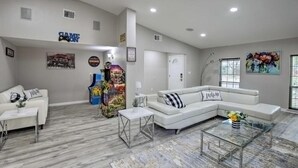 A spacious living room with a white sectional sofa, a glass coffee table, and a white couch. There are framed pictures on the walls, a game console sign, and a large window with blinds. - Collins Family-friendly Texas Home With Private Pool (Garland)