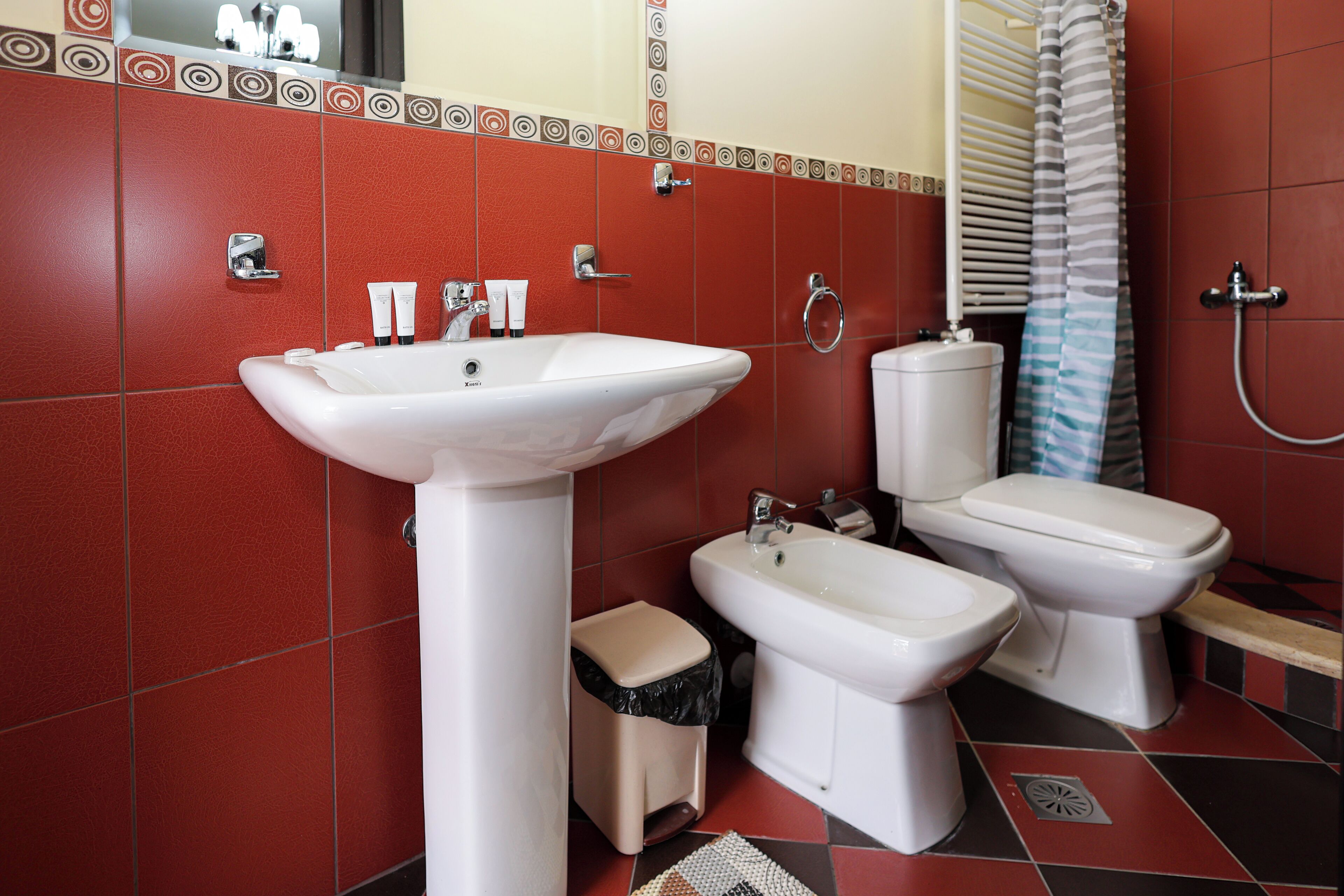 Une salle de bain avec des murs recouverts de carreaux rouges, un lavabo blanc, deux toilettes et un bidet.