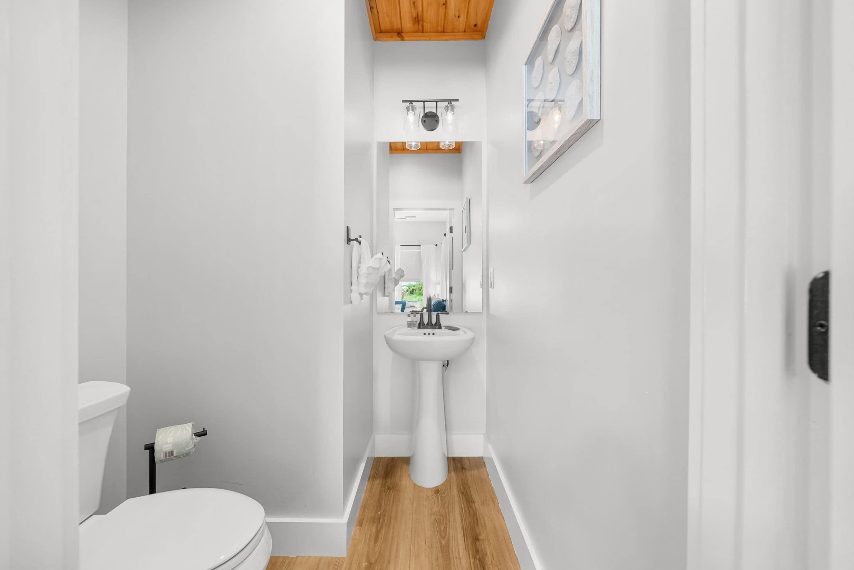 A narrow bathroom with a white sink, toilet, and wooden flooring.