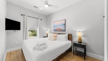 Un dormitorio con una cama grande, un ventilador de techo, un televisor de pantalla plana, un cuadro y una mesita de noche con lámpara.