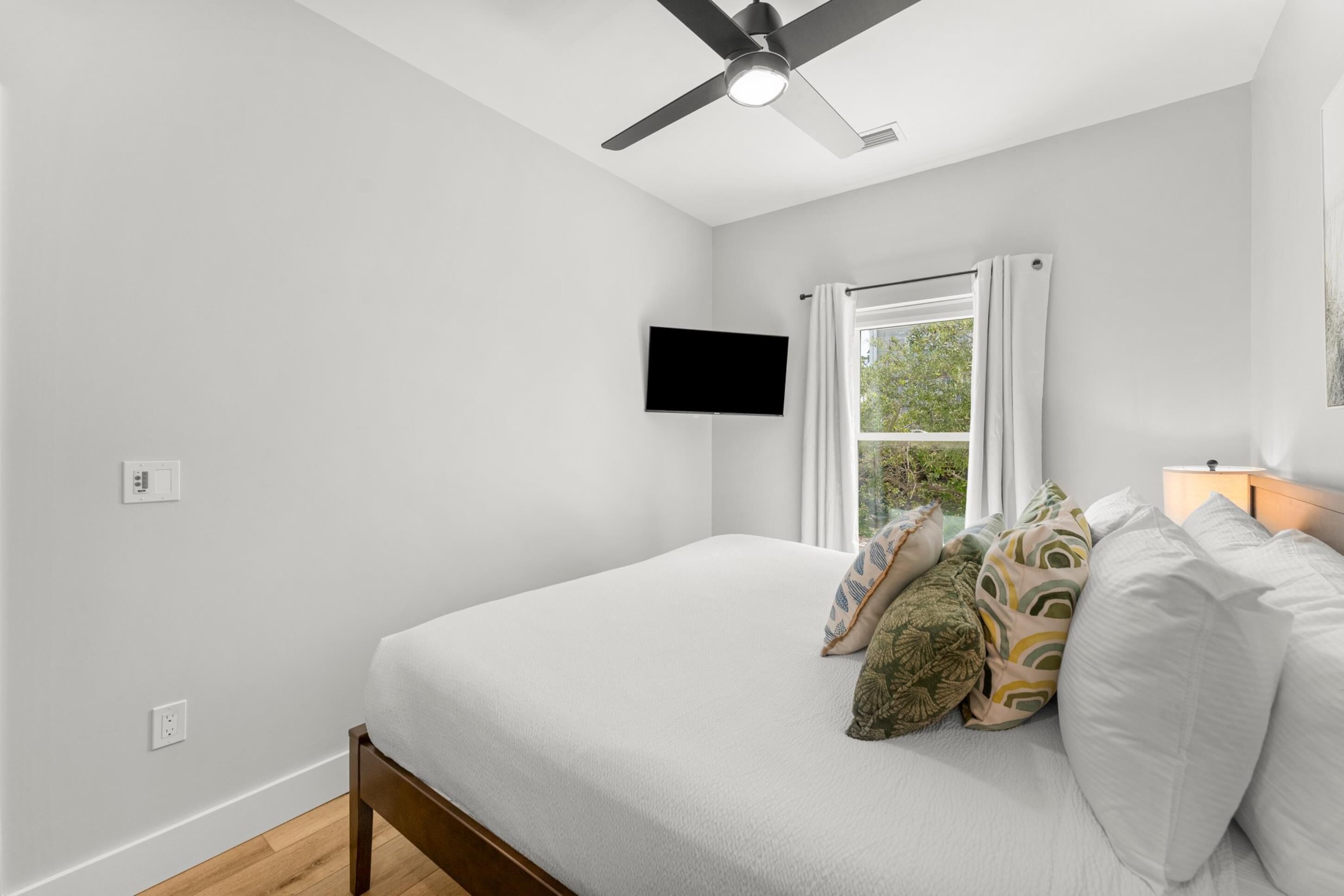A bedroom with a bed, a ceiling fan, a TV mounted on the wall, and a window with curtains.