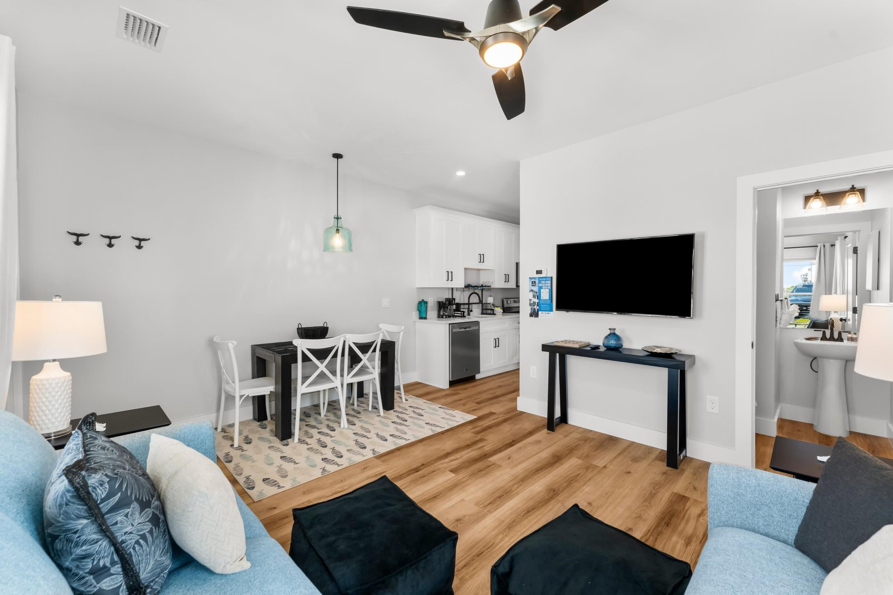A modern living room with a sofa, a coffee table, and a dining area with chairs and a table. There is a kitchen in the background with white cabinets and a TV mounted on the wall.
