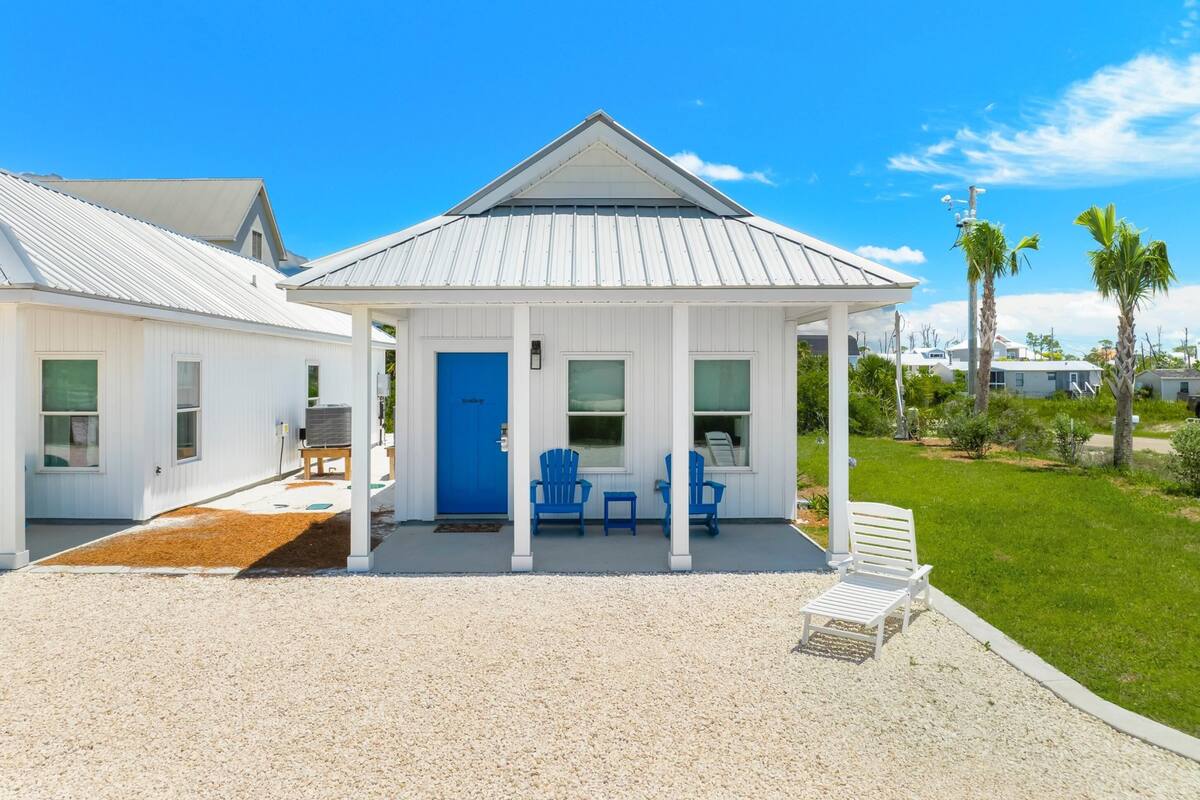 A two-story white building with a blue door and windows, a covered porch with blue chairs, and a gravel driveway.