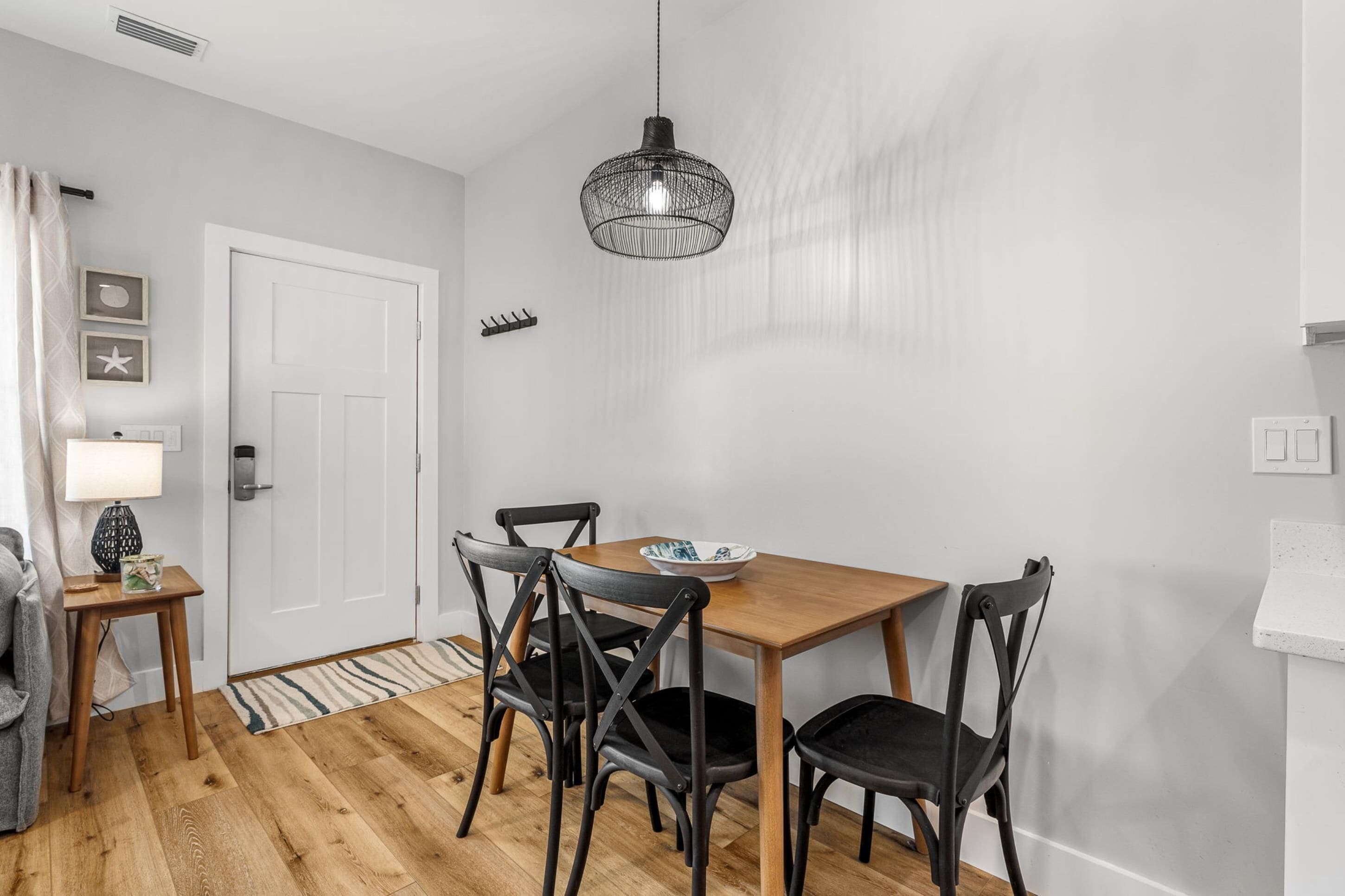 A dining area with a wooden table and four chairs, a hanging light fixture, and a white door.