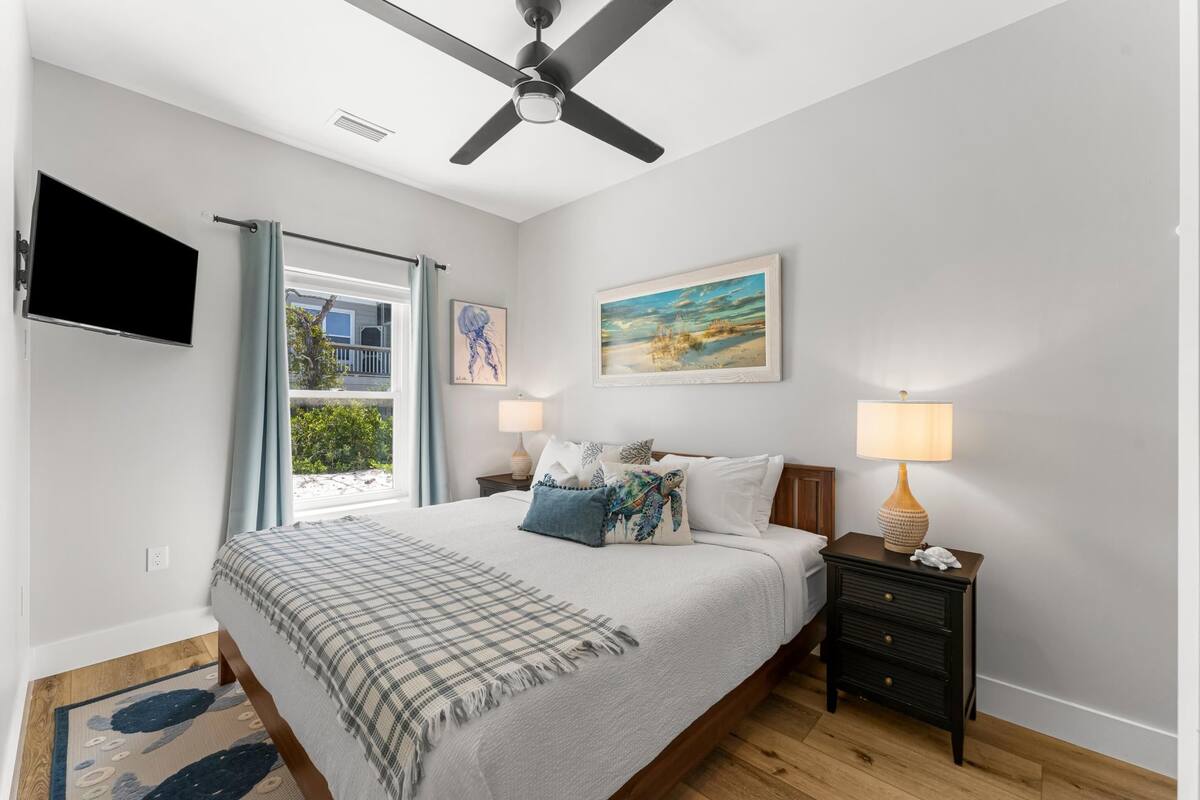 A bedroom with a bed, a ceiling fan, a TV, a window with curtains, a nightstand, and a lamp.