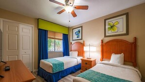 Une chambre d’hôtel avec deux lits, un bureau, un ventilateur de plafond, une fenêtre avec des rideaux et un tableau encadré au mur.