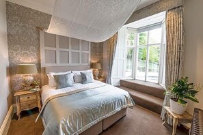 Makuuhuoneessa on suuri sänky, ikkunassa verhot, yöpöydällä lamppu ja ruukussa oleva huonekasvi.