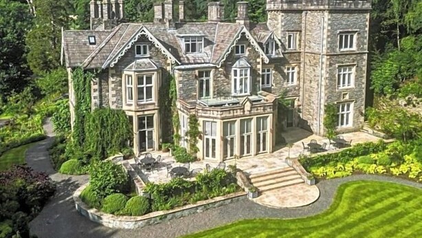 Un vaste manoir en pierre, doté de nombreuses cheminées, entouré d’une végétation luxuriante et d’une pelouse bien entretenue.
