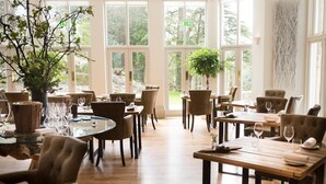 A spacious dining area with multiple tables and chairs, large windows with a view of greenery, and a central potted plant. - Forest Side Hotel (Ambleside)