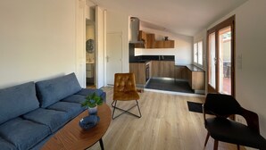 A modern living room with a sofa, a coffee table with a plant, and a chair. A kitchen with wooden cabinets and a sink is visible in the background. - The studio with its large, unoverlooked terrace, in a quiet location with private parking (Saint-Estève)